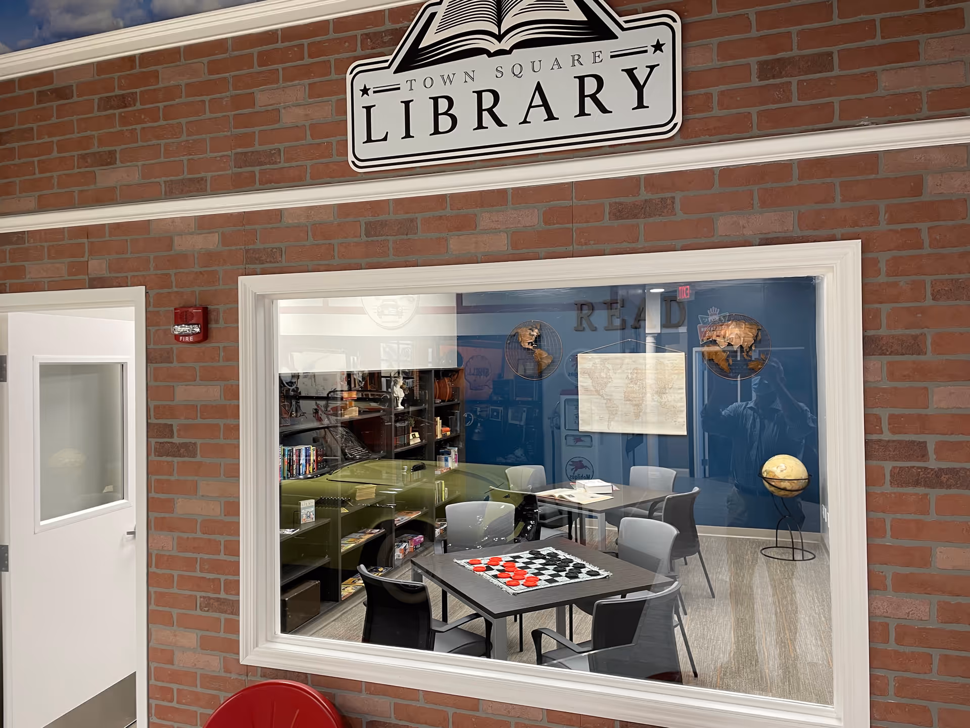 Brick exterior with large window revealing a library room with tables and shelves.