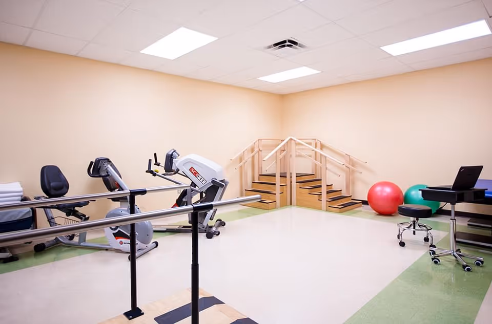 Indoor rehabilitation/exercise room with parallel bars, exercise bikes, a set of therapy stairs, exercise balls, and a rolling stool with a laptop on a table.