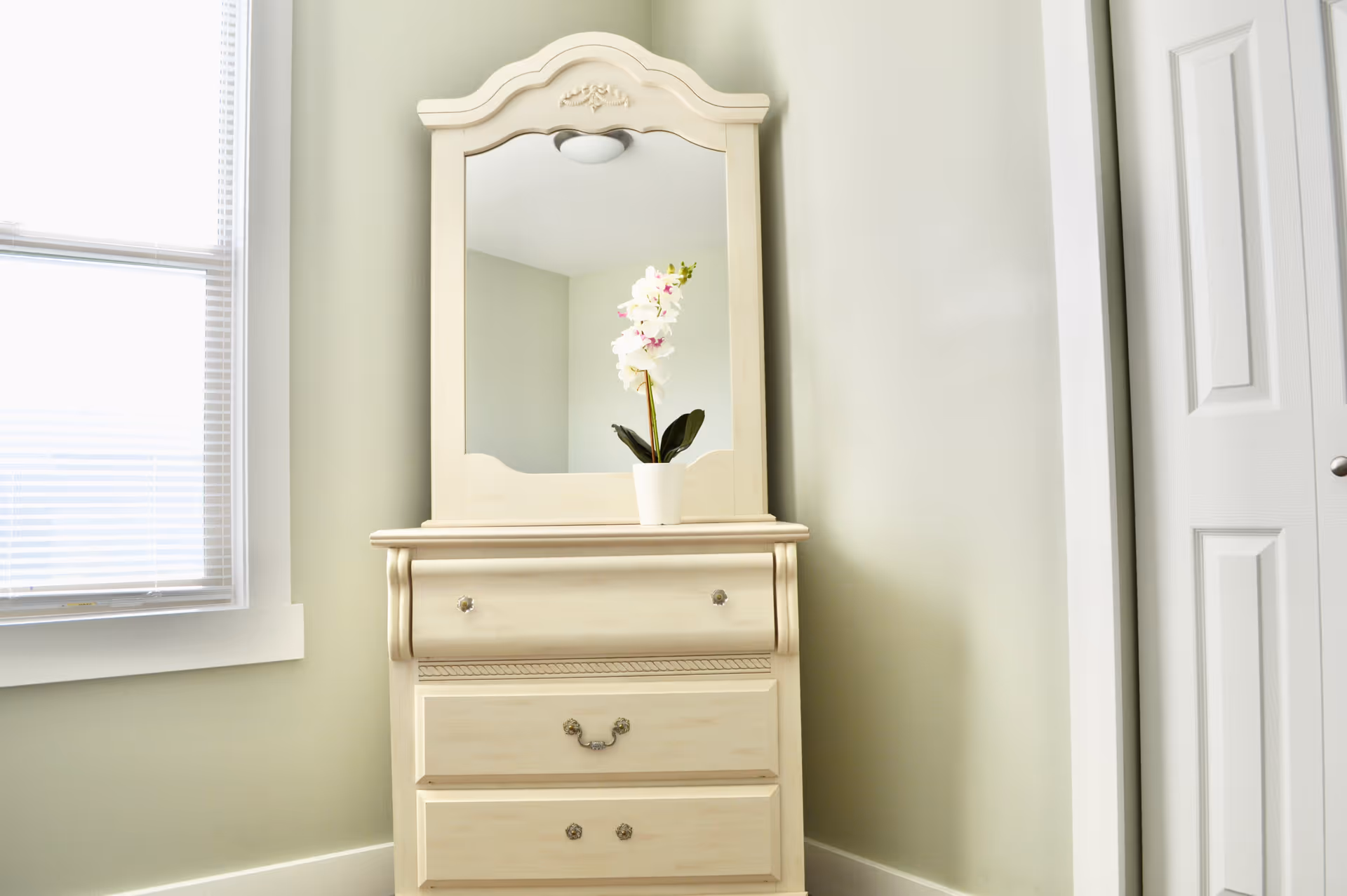 Cream dresser with mirror and orchid in a pale-green bedroom corner.