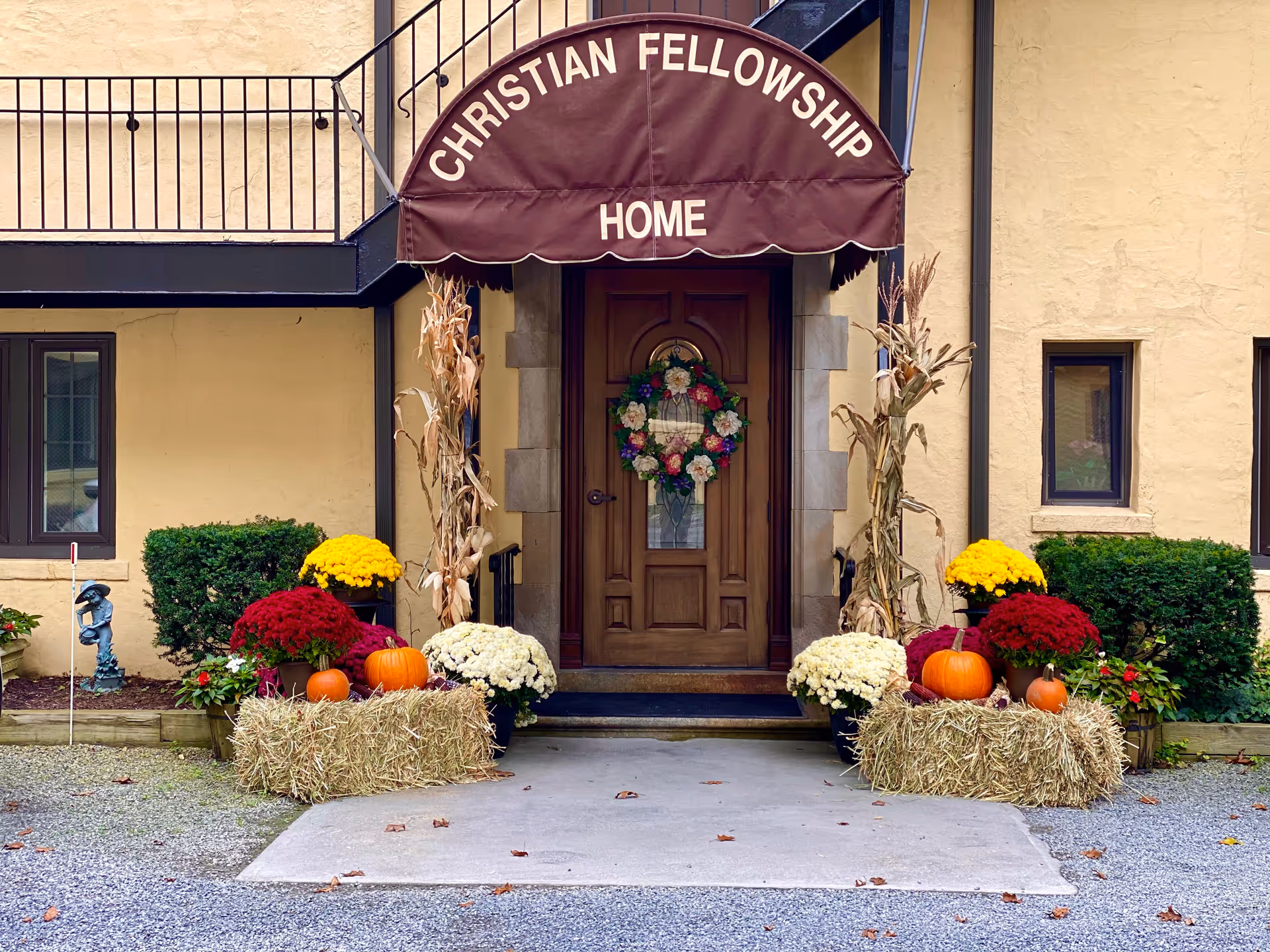 Entrance to Christian Fellowship Home with a brown awning displaying the facility name. The doorway is decorated with a floral wreath, and the entrance is flanked by hay bales adorned with pumpkins, red, yellow, and white flowers, and dried corn stalks. The building exterior is beige with two windows and some green bushes on either side.