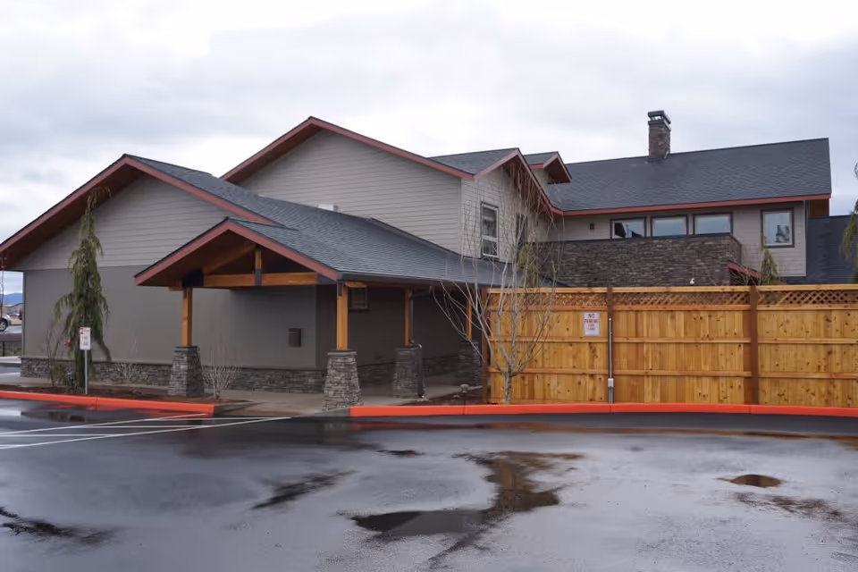 Exterior view of a modern building with gray siding, stone accents, and a dark shingled roof. The building has a covered entrance supported by stone pillars and wooden beams. A wooden fence is visible on the right side, and the ground is wet with puddles, indicating recent rain. The sky is overcast.