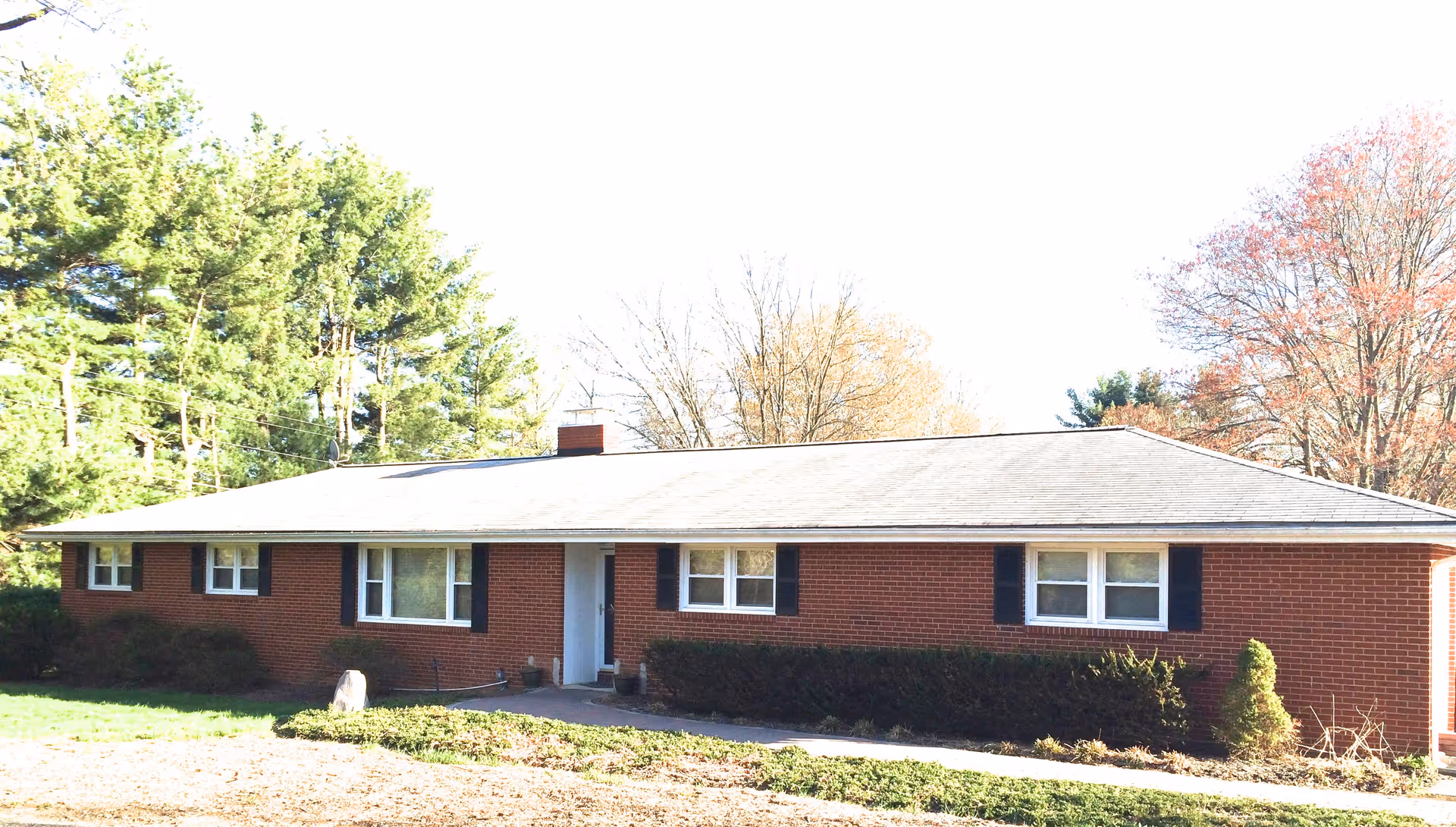 Front view of a single-story red brick building with white-trimmed windows, a centered entrance, shrubs, and trees under a bright sky.