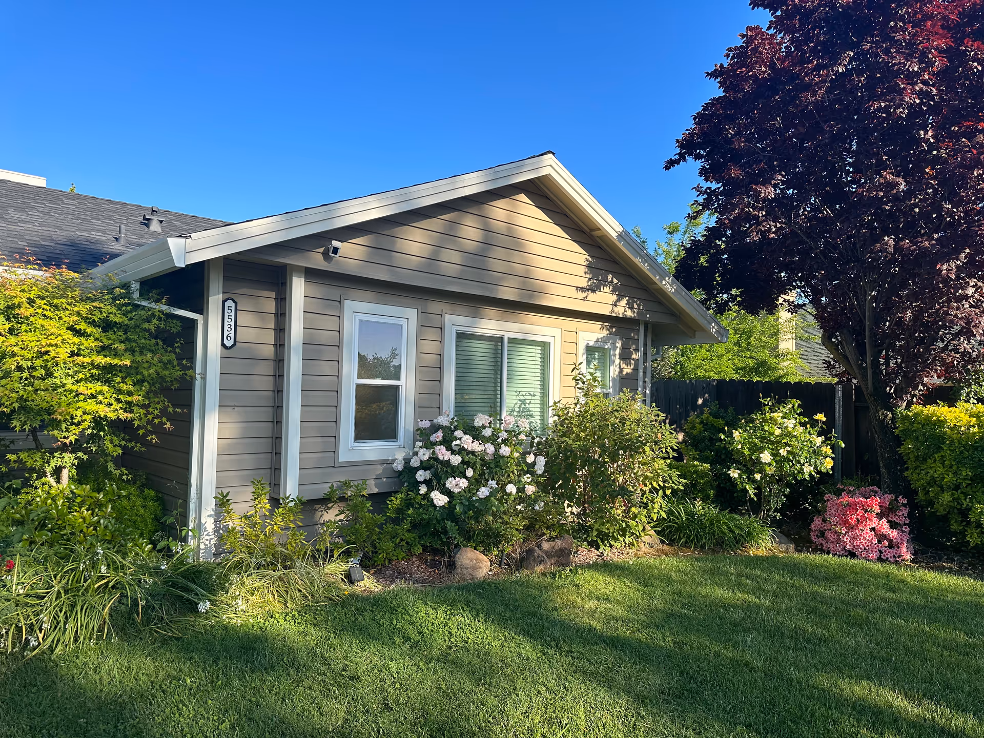 A single-story beige house with a well-kept lawn and flowering shrubs in front.