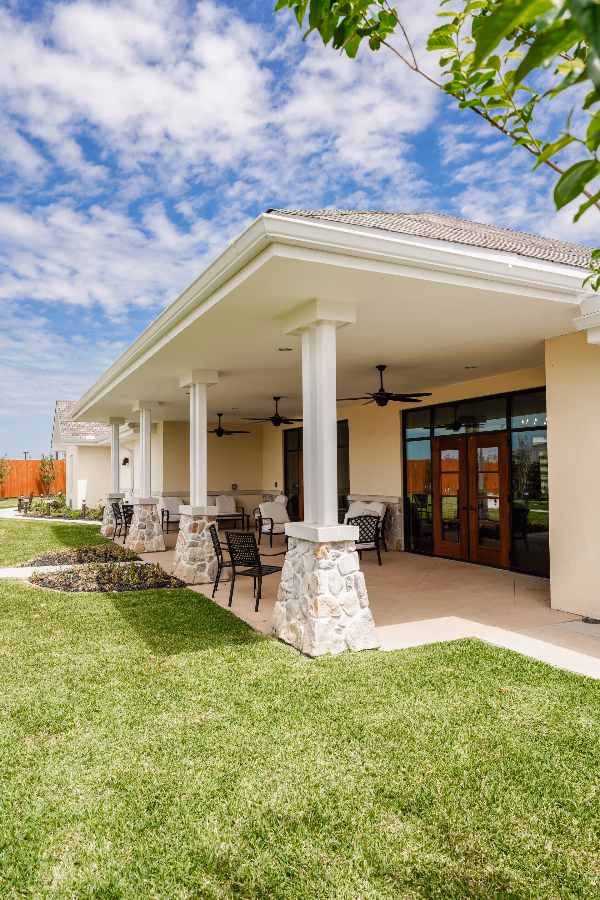 Outdoor covered patio area with stone pillars and ceiling fans at The Harbor Assisted Living, featuring several chairs and tables, surrounded by green grass and a partly cloudy sky.