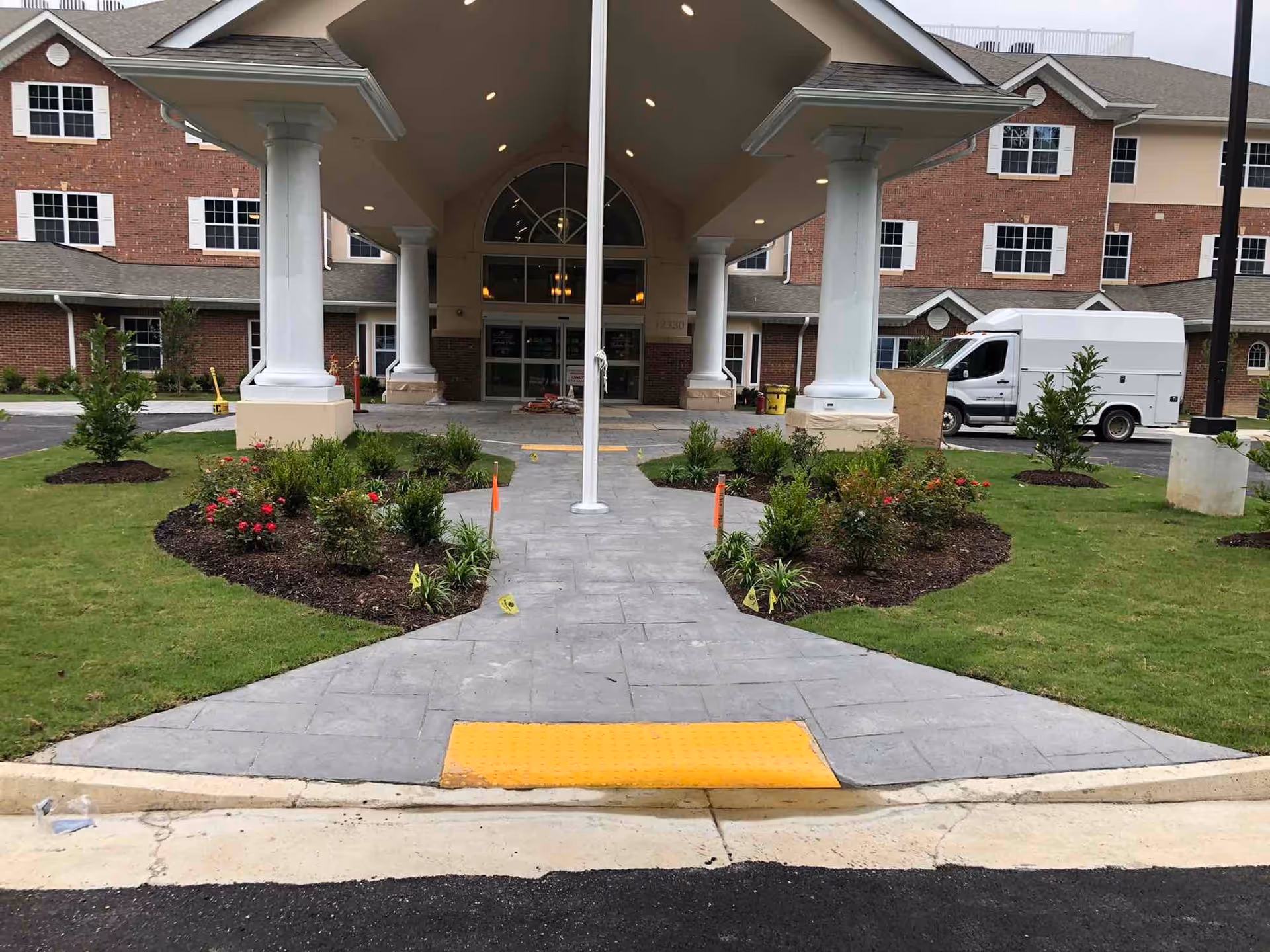Covered main entrance to a brick retirement community building with a paved walkway, large white columns, landscaping and a white service van.