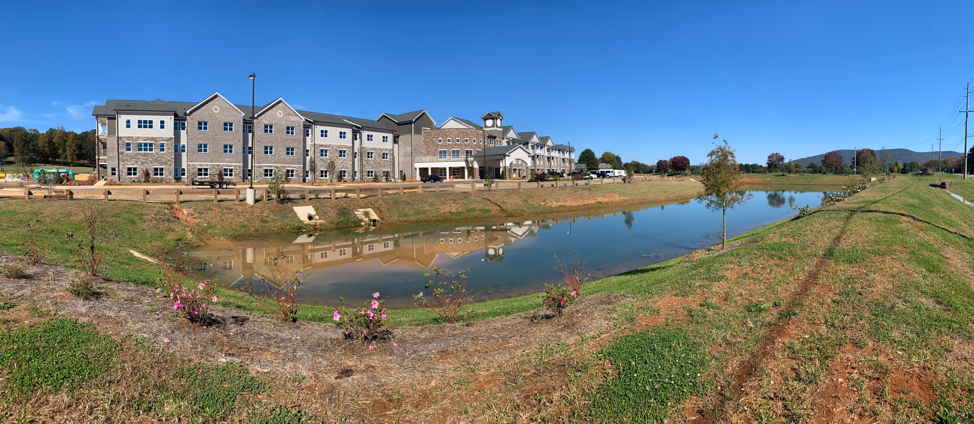 Panoramic view of a multi-story senior living building reflected in a pond with grassy landscaping under a clear blue sky.