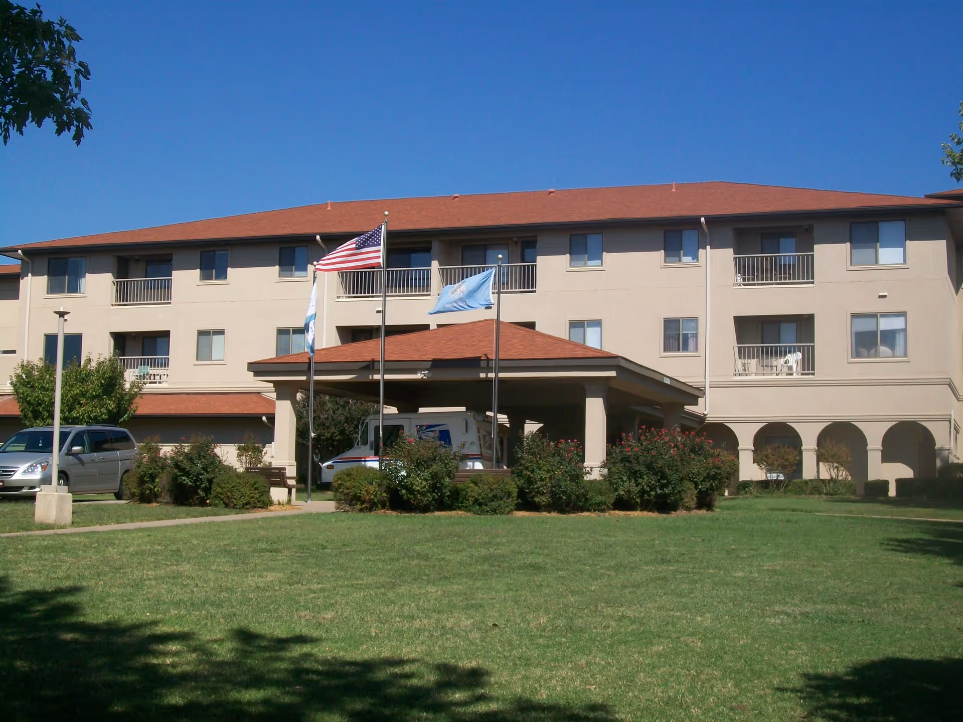 Exterior view of a three-story senior living facility building with a red roof and beige walls. There are balconies on the second and third floors, a covered entrance with three flagpoles displaying the American flag and two other flags, and a green lawn with bushes in front. A USPS mail truck and a silver minivan are parked near the entrance.