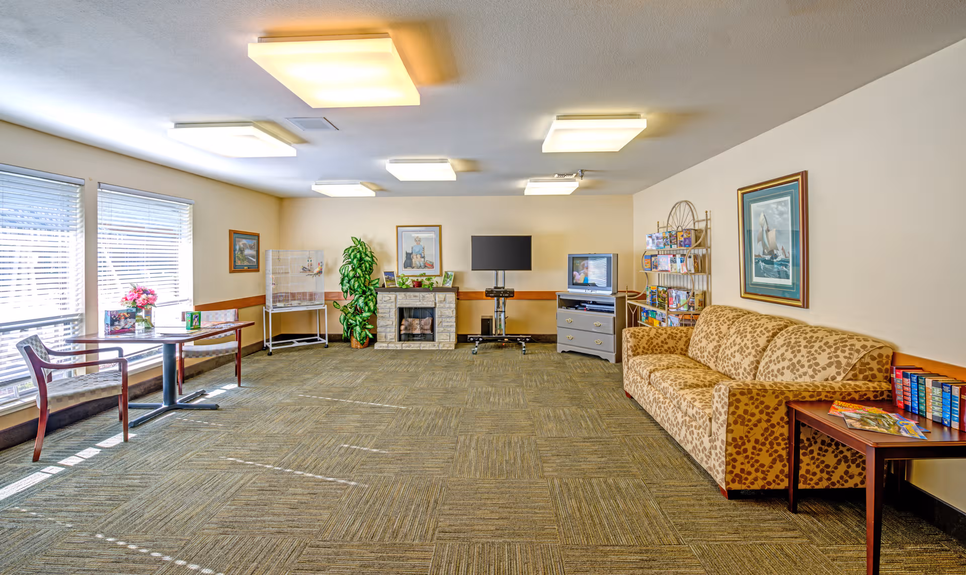Bright senior living lounge with sofa, tables, TV, magazines, and large windows.