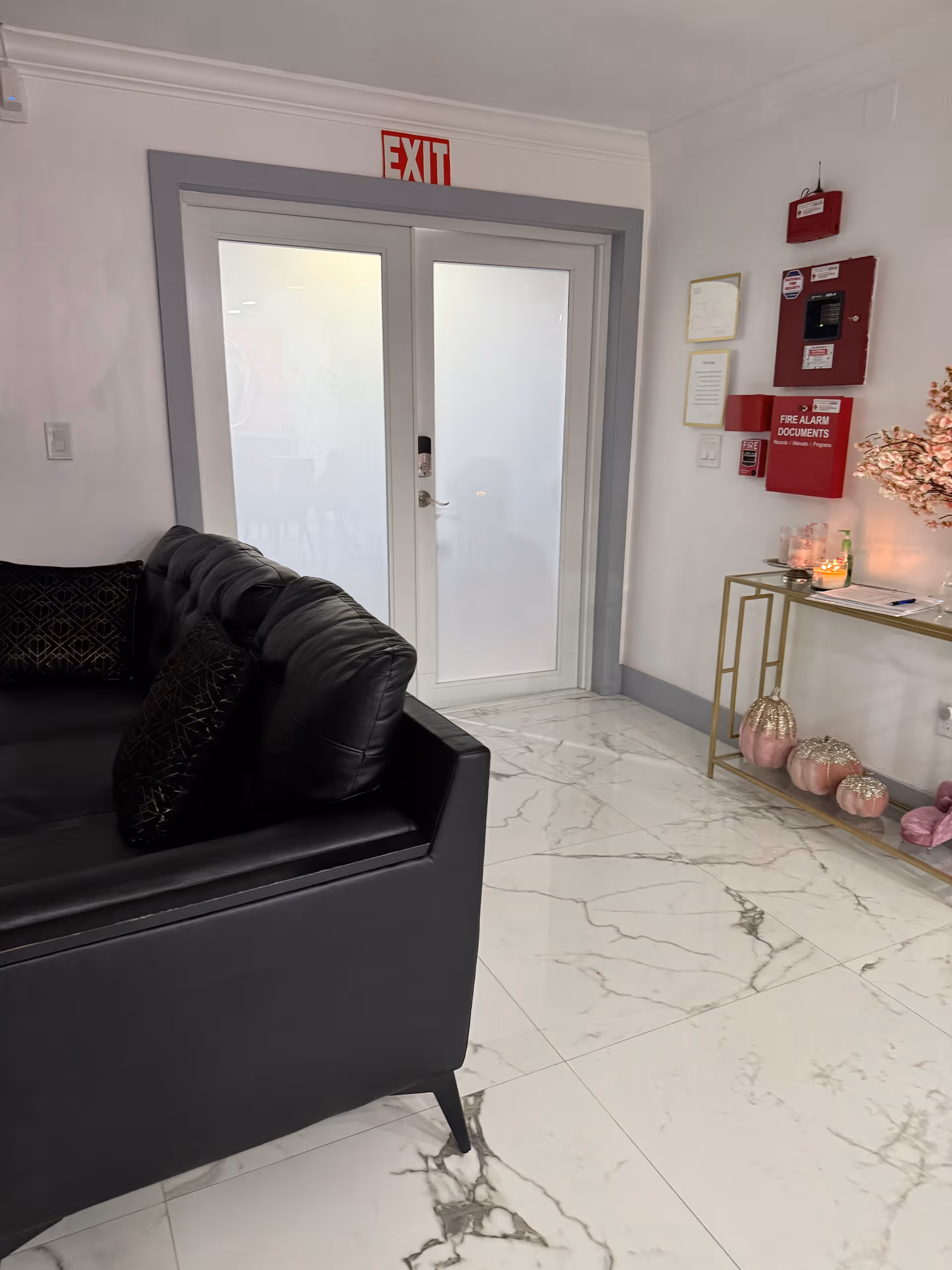 Modern lobby seating area with black sofa, marble floor, glass exit doors, red fire equipment.