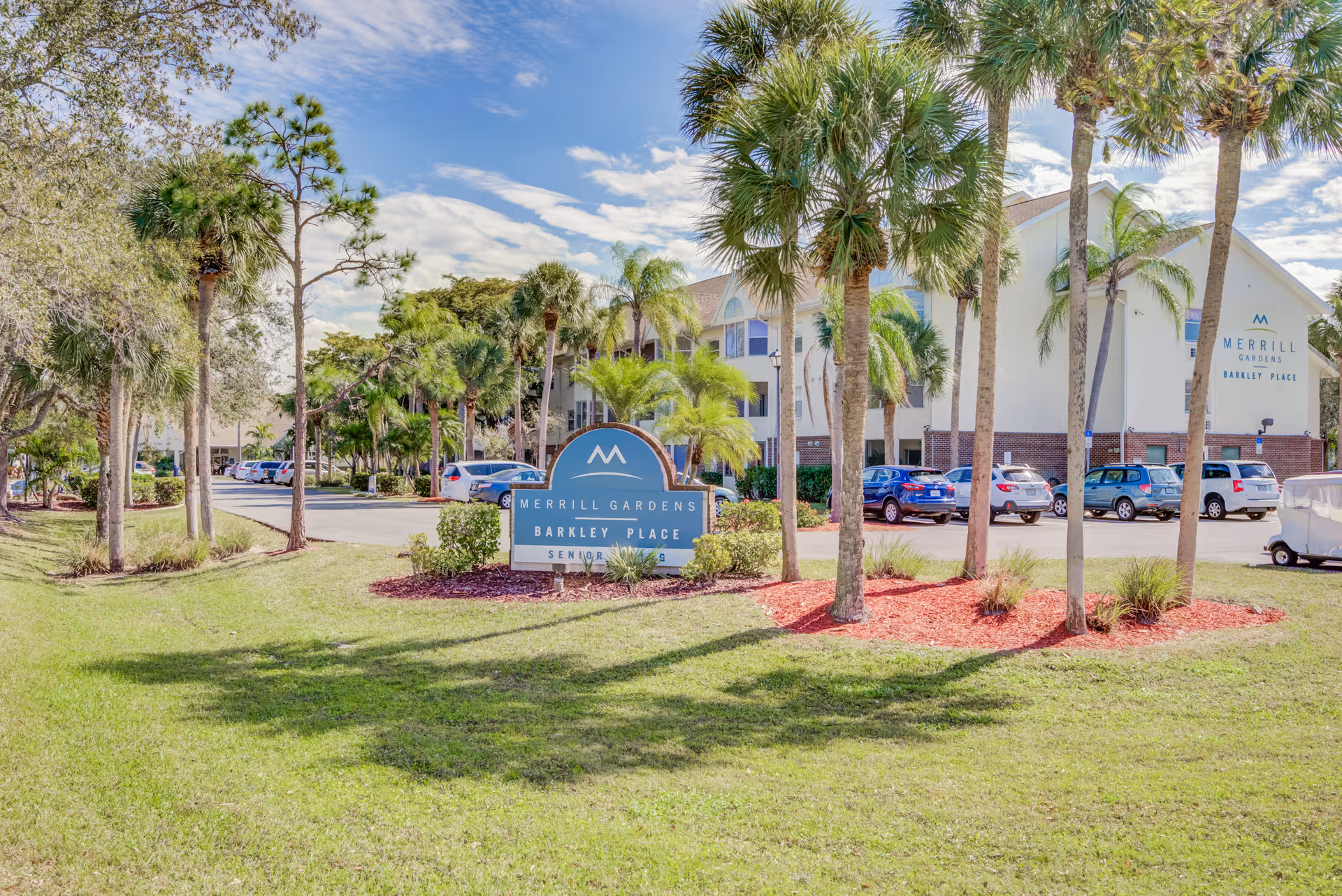 Exterior view of Merrill Gardens at Barkley Place with a sign, palm trees, and parked cars.