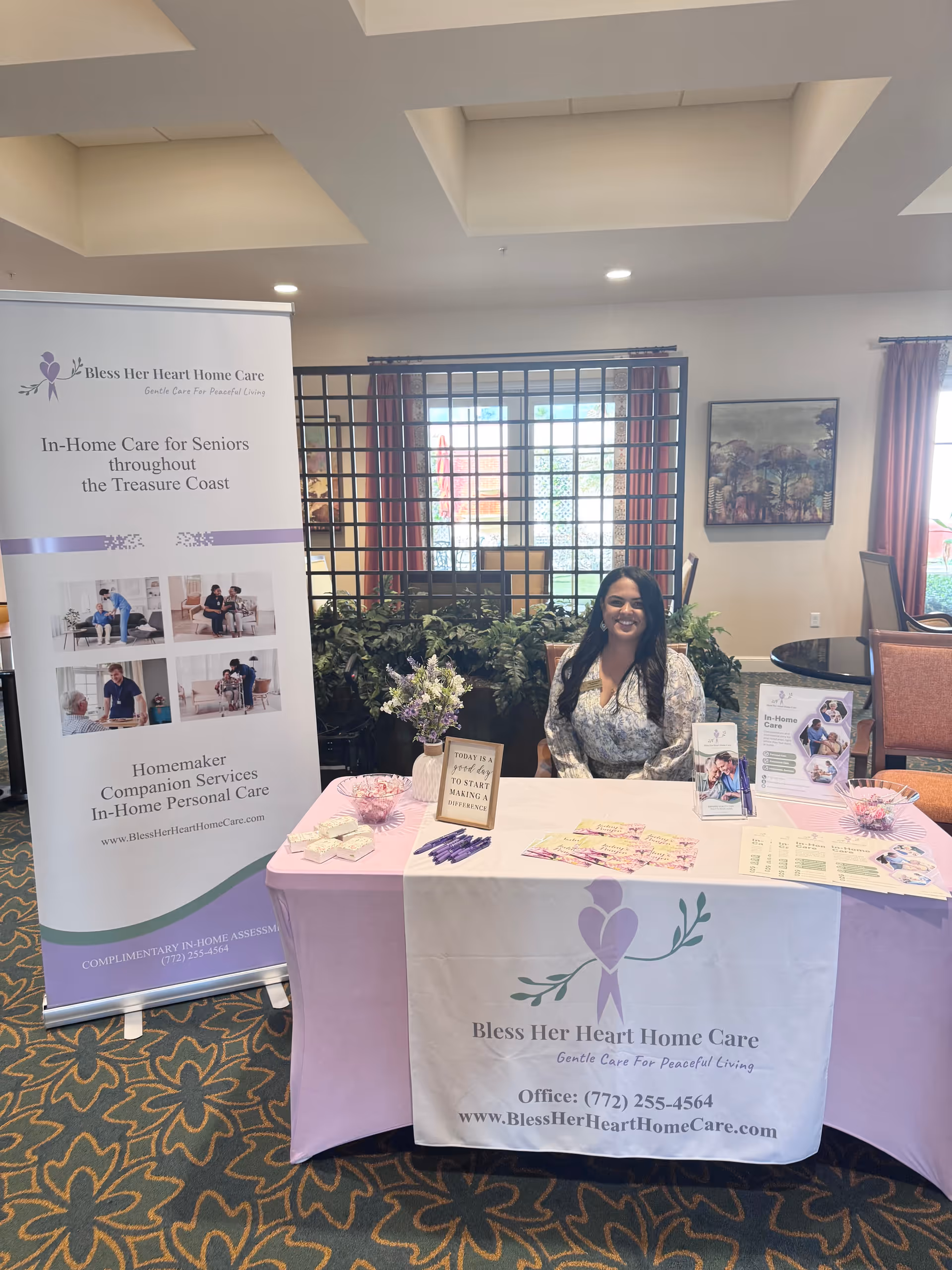 Smiling woman sits at a promotional table with banners for Bless Her Heart Home Care indoors.