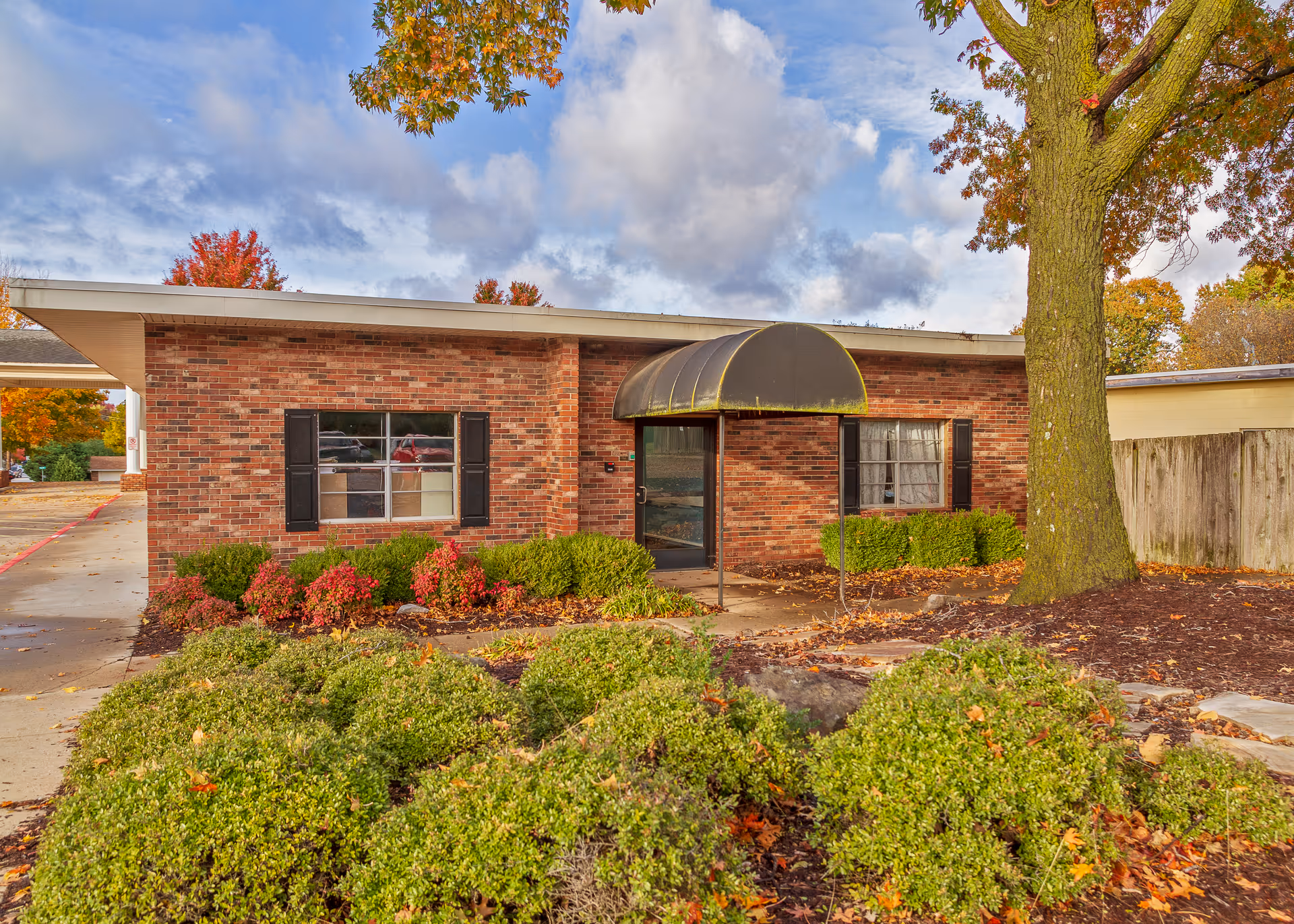 Exterior view of a single-story brick building with black shutters on the windows and a black awning over the entrance door. The building is surrounded by green bushes, a large tree, and a concrete walkway. The sky is partly cloudy with autumn foliage visible on the trees.