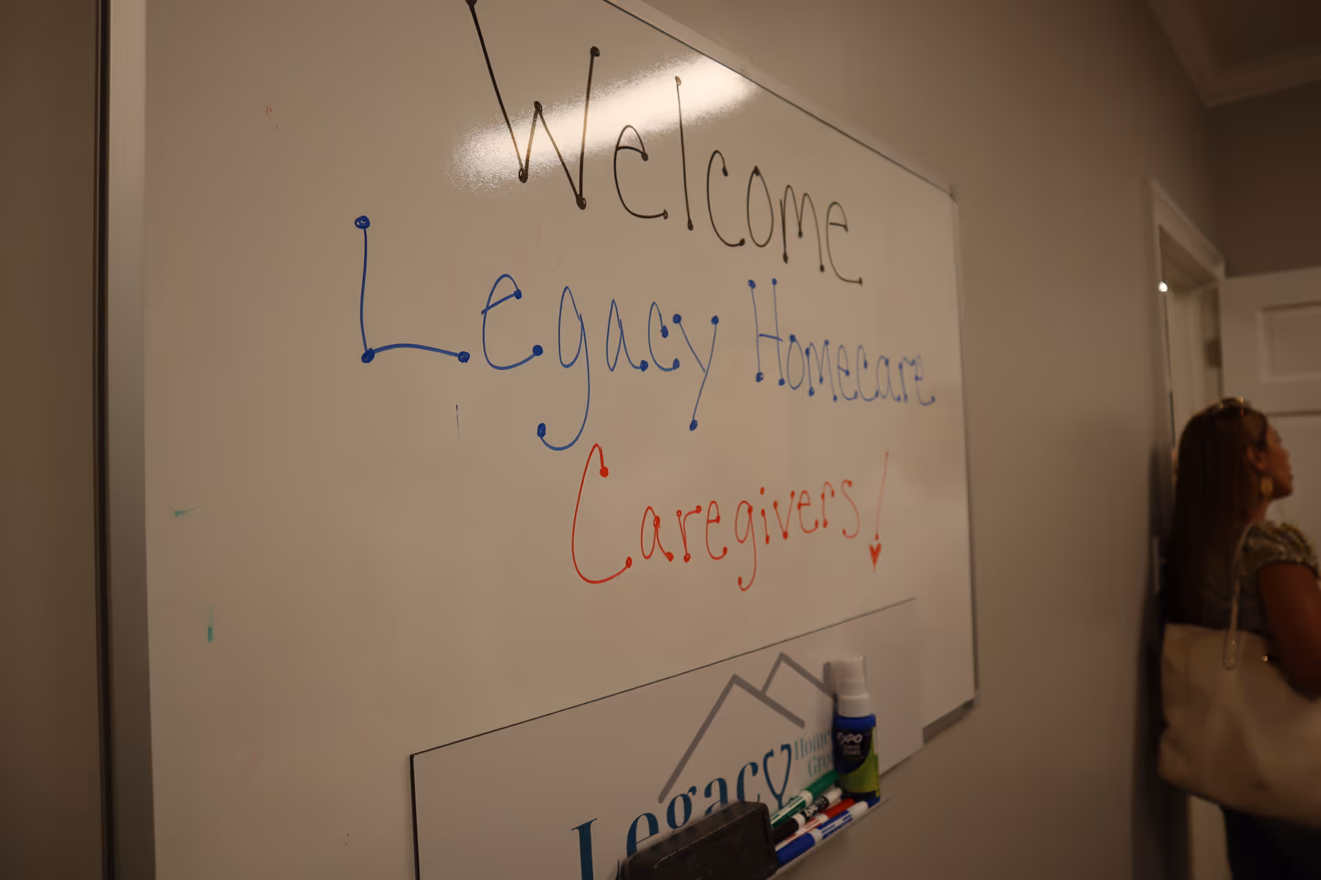 Whiteboard shows Welcome Legacy Homecare Caregivers in a hallway beside a standing woman.