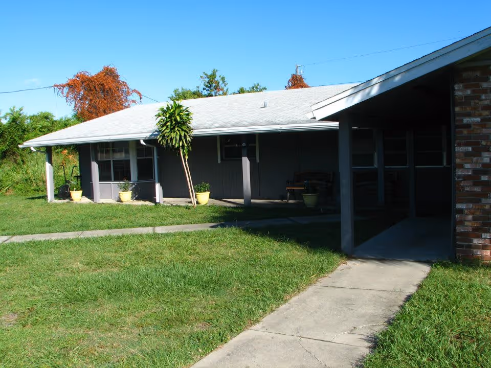 Exterior view of a single-story assisted living facility building with a covered porch, several potted plants, a concrete walkway, and a grassy lawn under a clear blue sky.