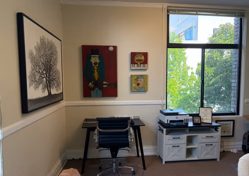 A small office corner with desk, chair, printer, and wall art near a large window.