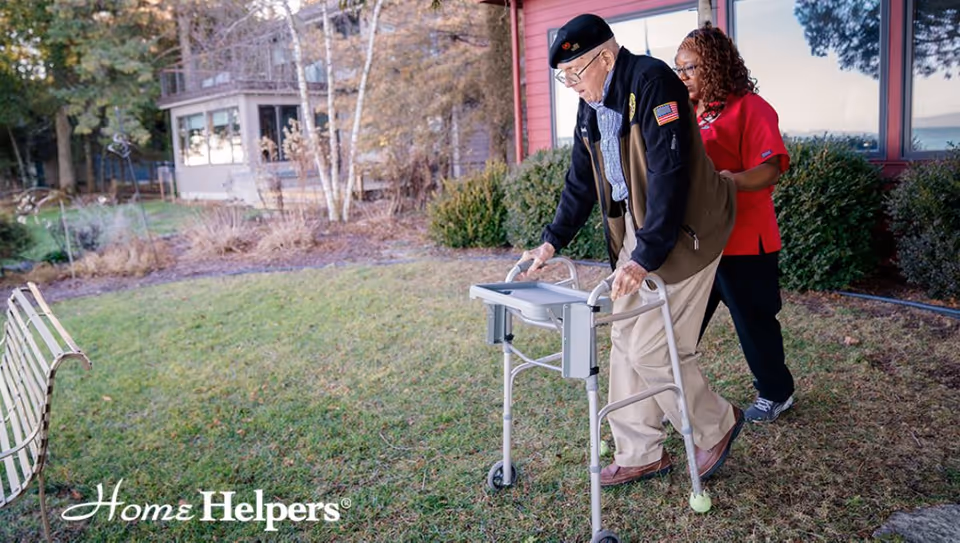 An elderly man with a walker aided by a caregiver outdoors near a red building