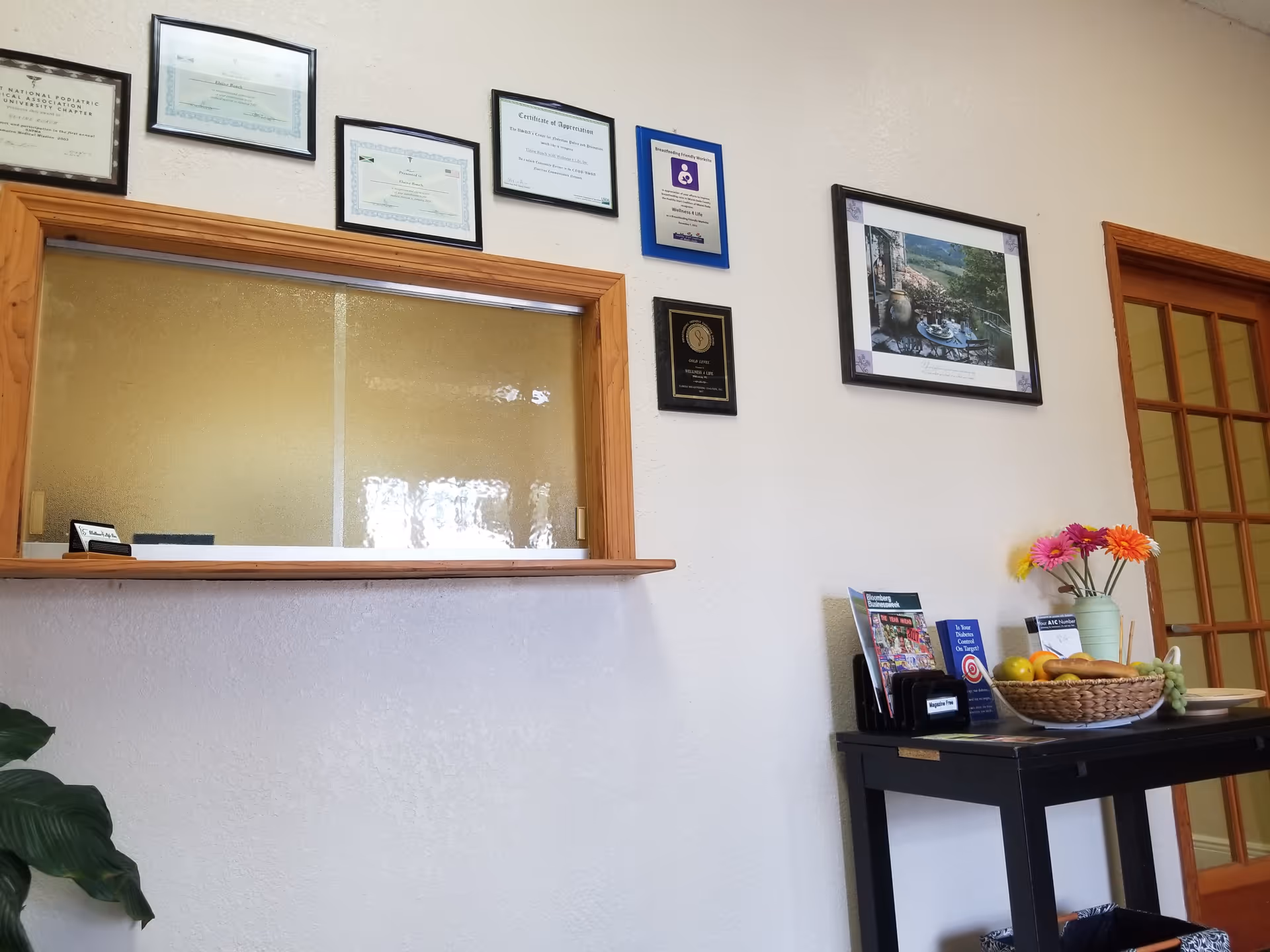 Reception window with framed certificates and a table holding a fruit basket and flowers.