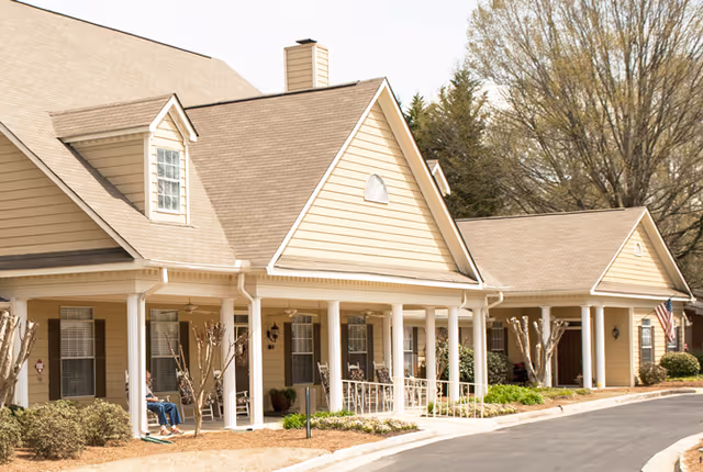 Exterior view of a beige senior living facility building with a covered porch supported by white columns. Several rocking chairs are placed on the porch, and there are small bushes and trees in front of the building. An American flag is visible near the entrance, and the driveway curves along the front of the building.