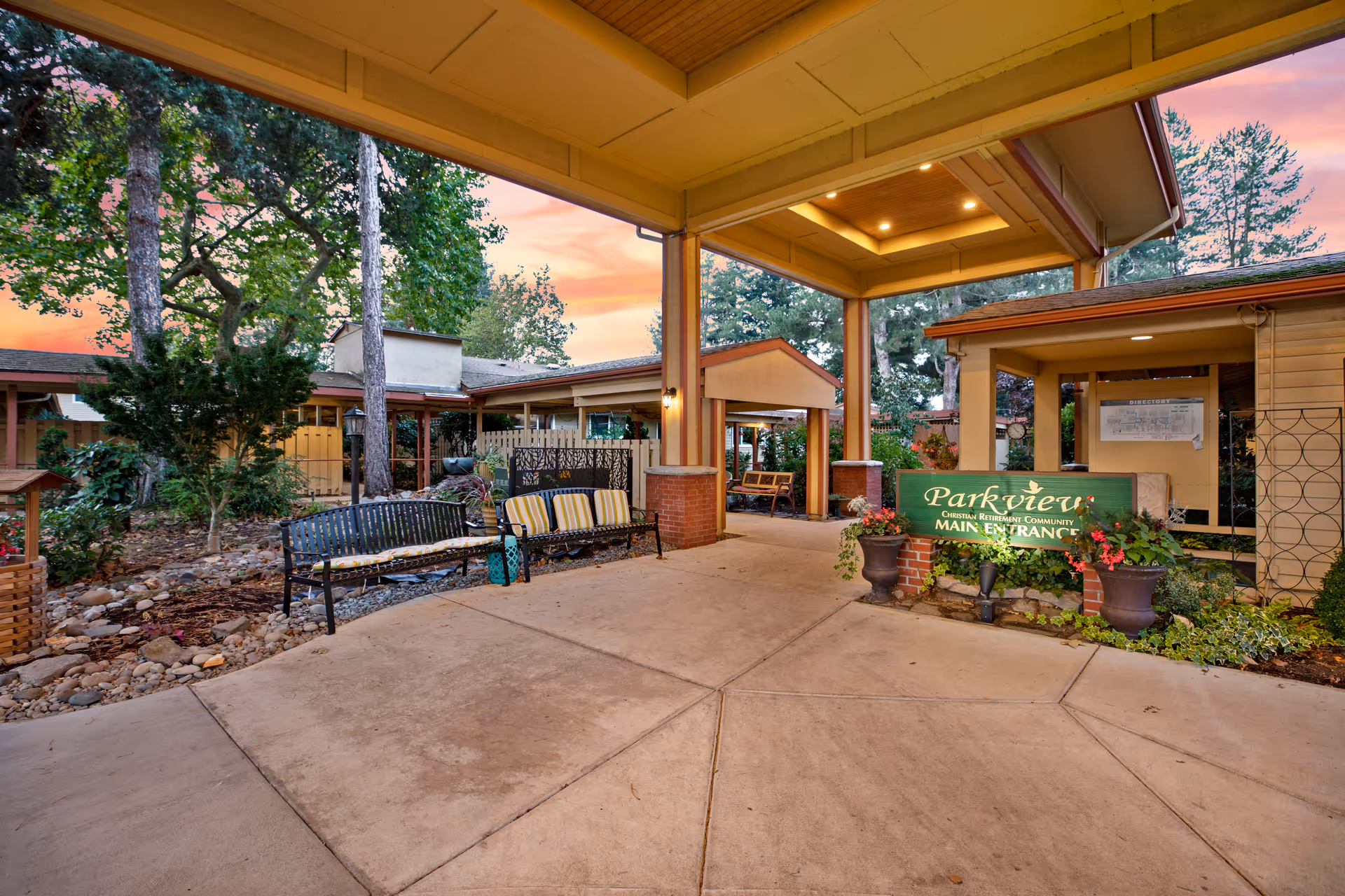 Covered entrance area of Parkview Christian Retirement Community at sunset, featuring benches with cushions, potted plants, trees, and a green sign indicating the main entrance.