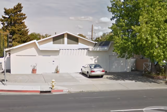 White mid-century building front with a parked car and green trees along a quiet street.
