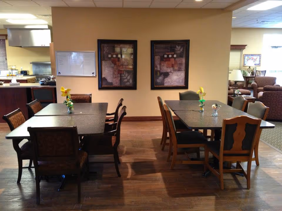Interior view of a dining area in Lakewood Assisted Living with two rectangular tables, each surrounded by six chairs. Each table has a small vase with yellow flowers. The room has wooden flooring, beige walls with two framed abstract paintings, and an adjacent kitchen area visible on the left. In the background, there is a lounge area with armchairs and a lamp.