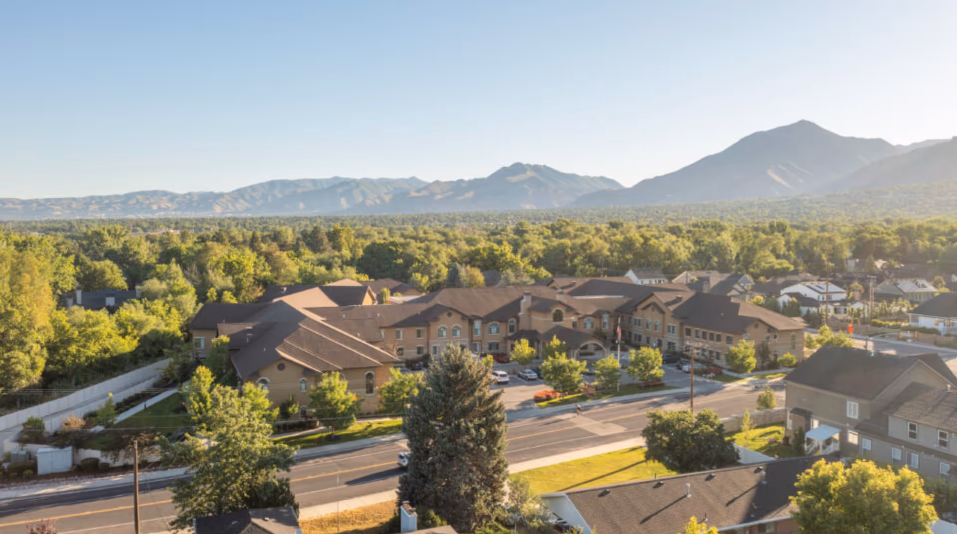 Aerial view of a senior living complex with multiple connected buildings, a tree-lined road in front, and mountains in the distance.