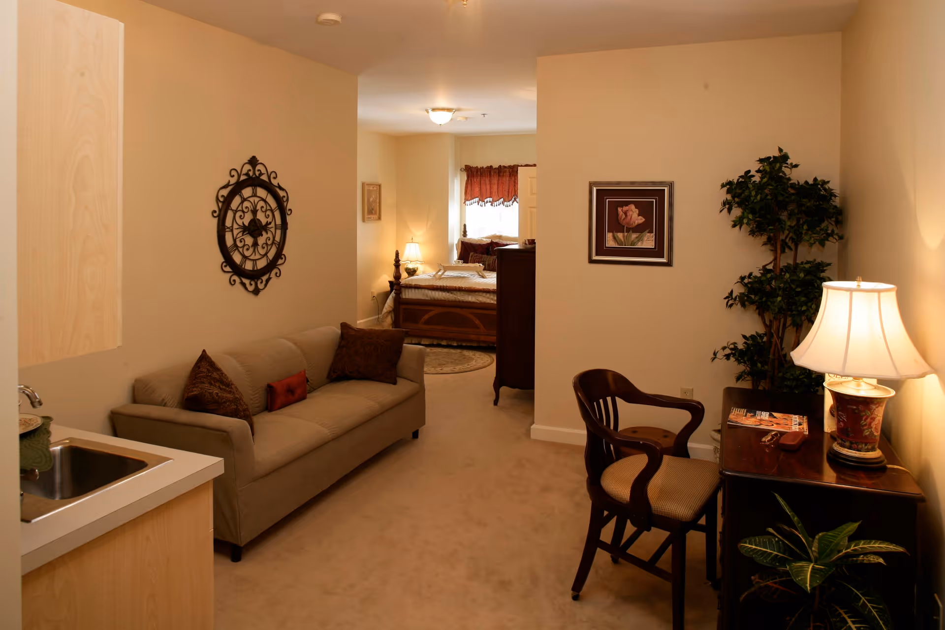 Interior view of a senior living facility room featuring a beige sofa with pillows, a small kitchenette with a sink, a wooden chair, a desk with a lamp and magazines, a potted plant, and a bedroom area visible in the background with a bed, nightstand, and lamp.