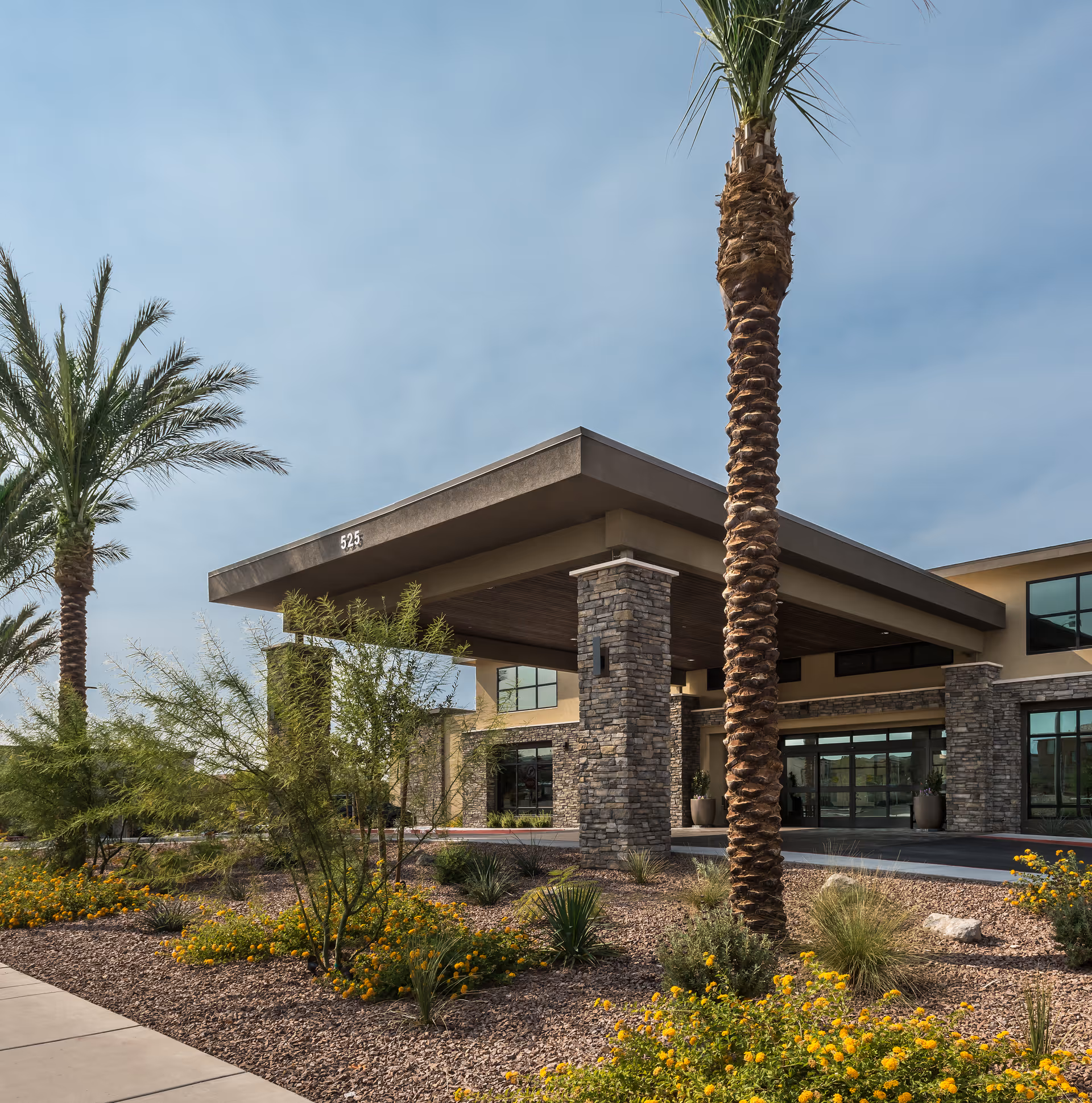 Exterior view of Carnegie Heights at Henderson facility showing the entrance with stone pillars, a covered drop-off area, palm trees, and landscaped garden with yellow flowers under a clear sky.