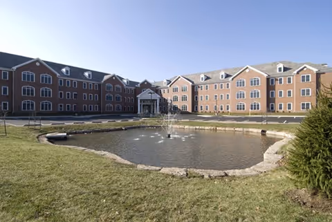 Brick senior living building with a pond and fountain and a grassy lawn in front under a clear sky.