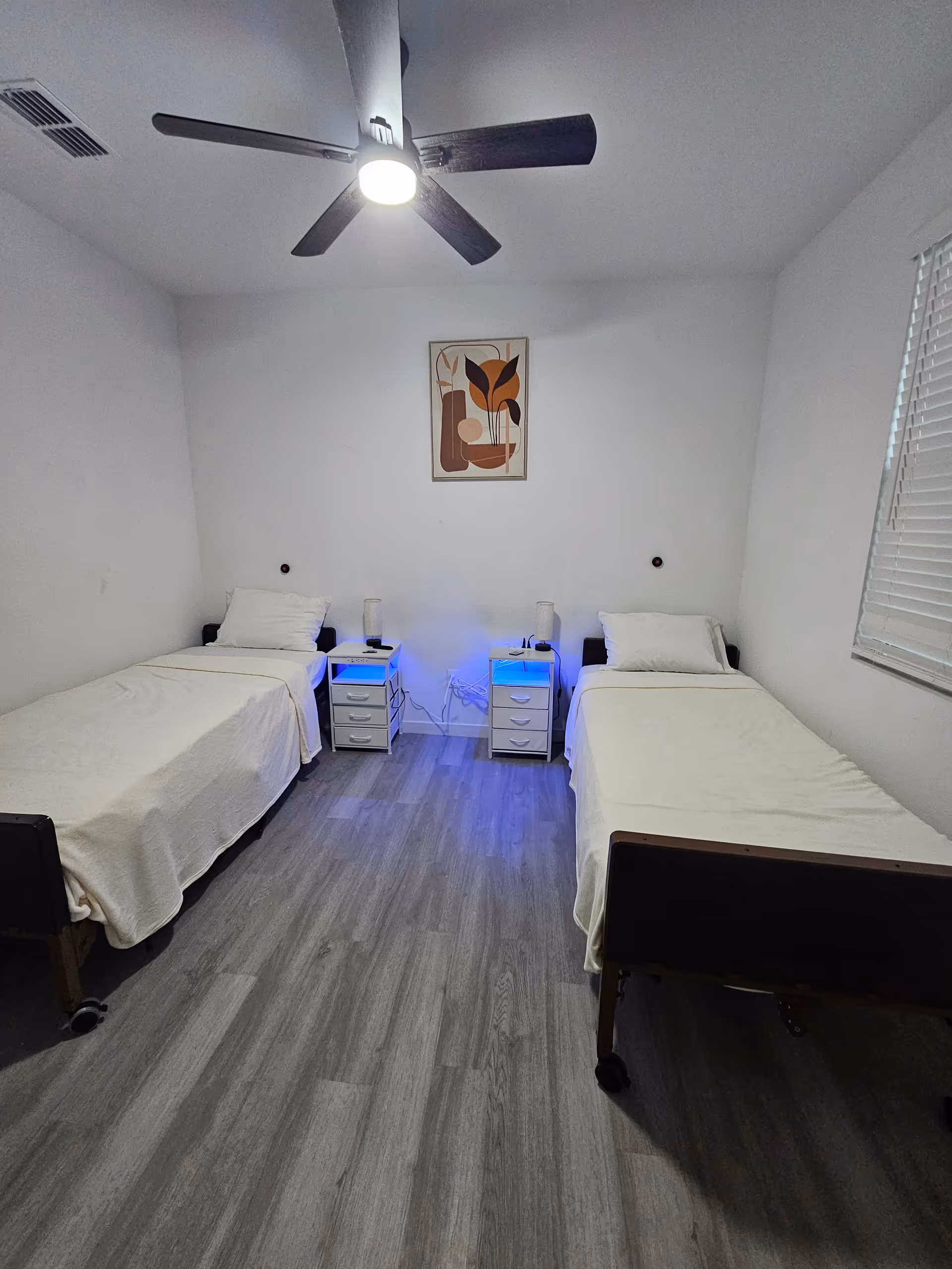 A simple bedroom with two single beds placed parallel to each other against opposite walls. Each bed has a white pillow and beige blanket. Between the beds are two small white nightstands with blue LED lights underneath, each holding a lamp and some small items. A modern ceiling fan with a light is mounted on the ceiling. A framed abstract artwork hangs on the wall above the nightstands. The floor is covered with gray wood-like vinyl planks, and there is a window with white blinds on the right wall.