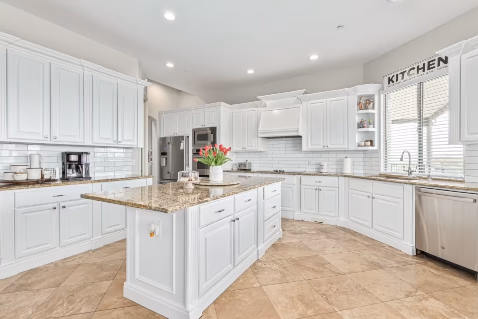 Bright and spacious kitchen with white cabinetry, granite countertops, and a large central island. The kitchen features stainless steel appliances including a refrigerator, oven, and dishwasher. There is a window with blinds above the sink and a decorative sign that reads 'KITCHEN' on the wall. The floor is tiled with beige tiles, and there are recessed lights in the ceiling.