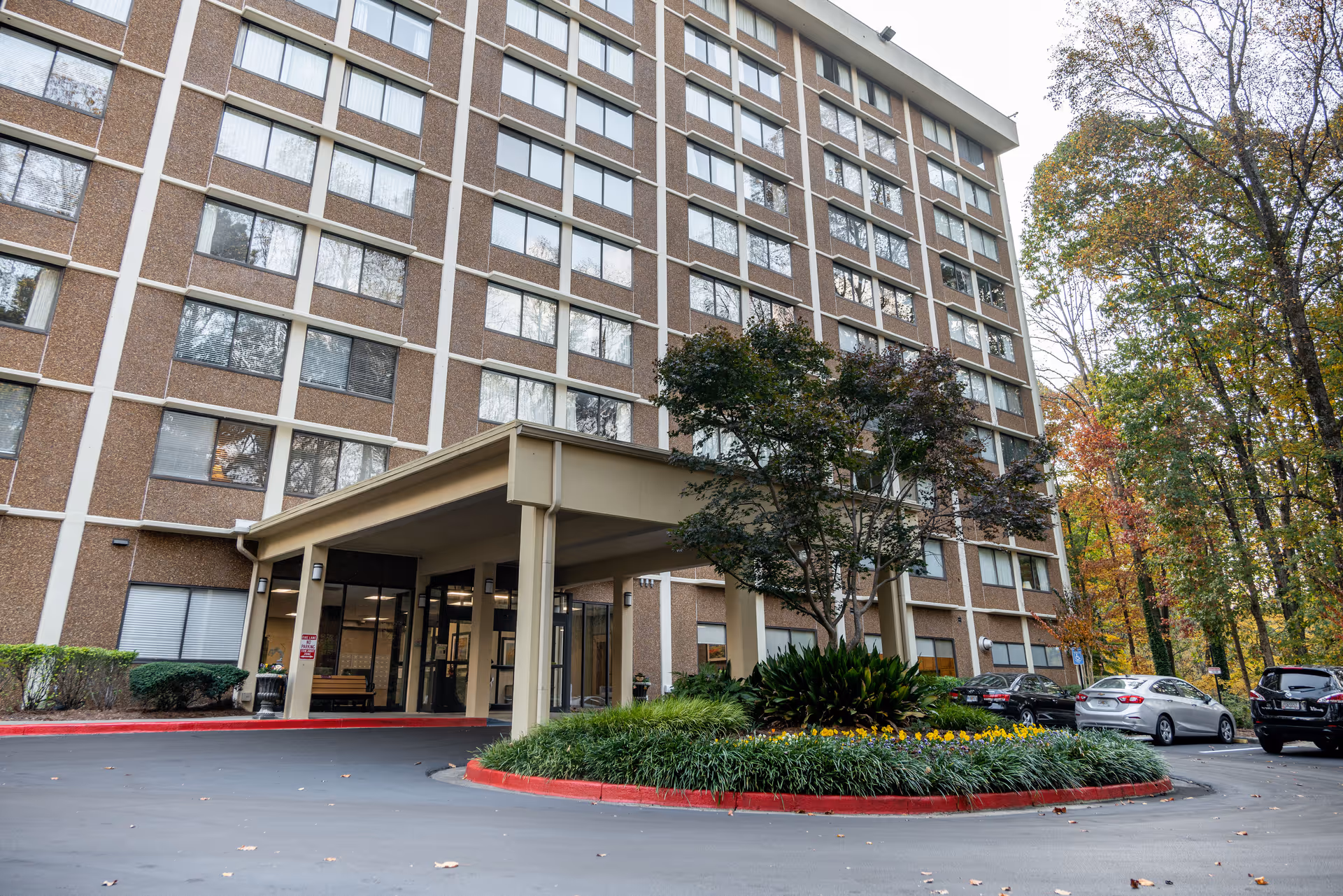 Exterior view of a multi-story senior apartment building with a covered entrance and a circular driveway surrounded by landscaped greenery and trees with autumn foliage. Several cars are parked near the entrance.
