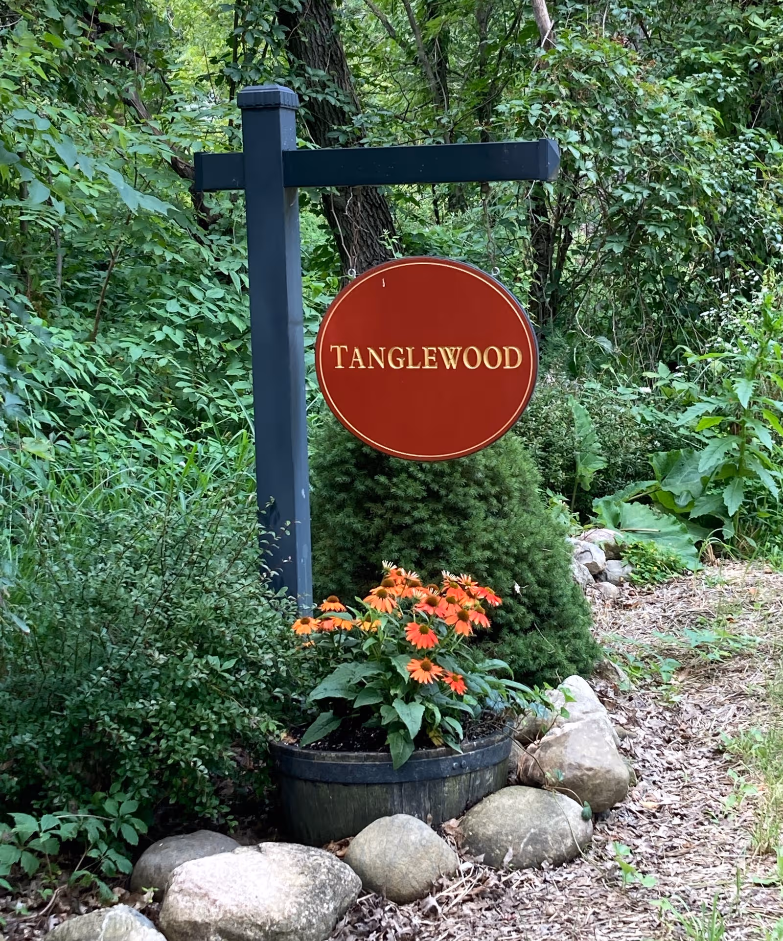 A red circular Tanglewood sign hangs from a dark blue post amid lush greenery and rocks.