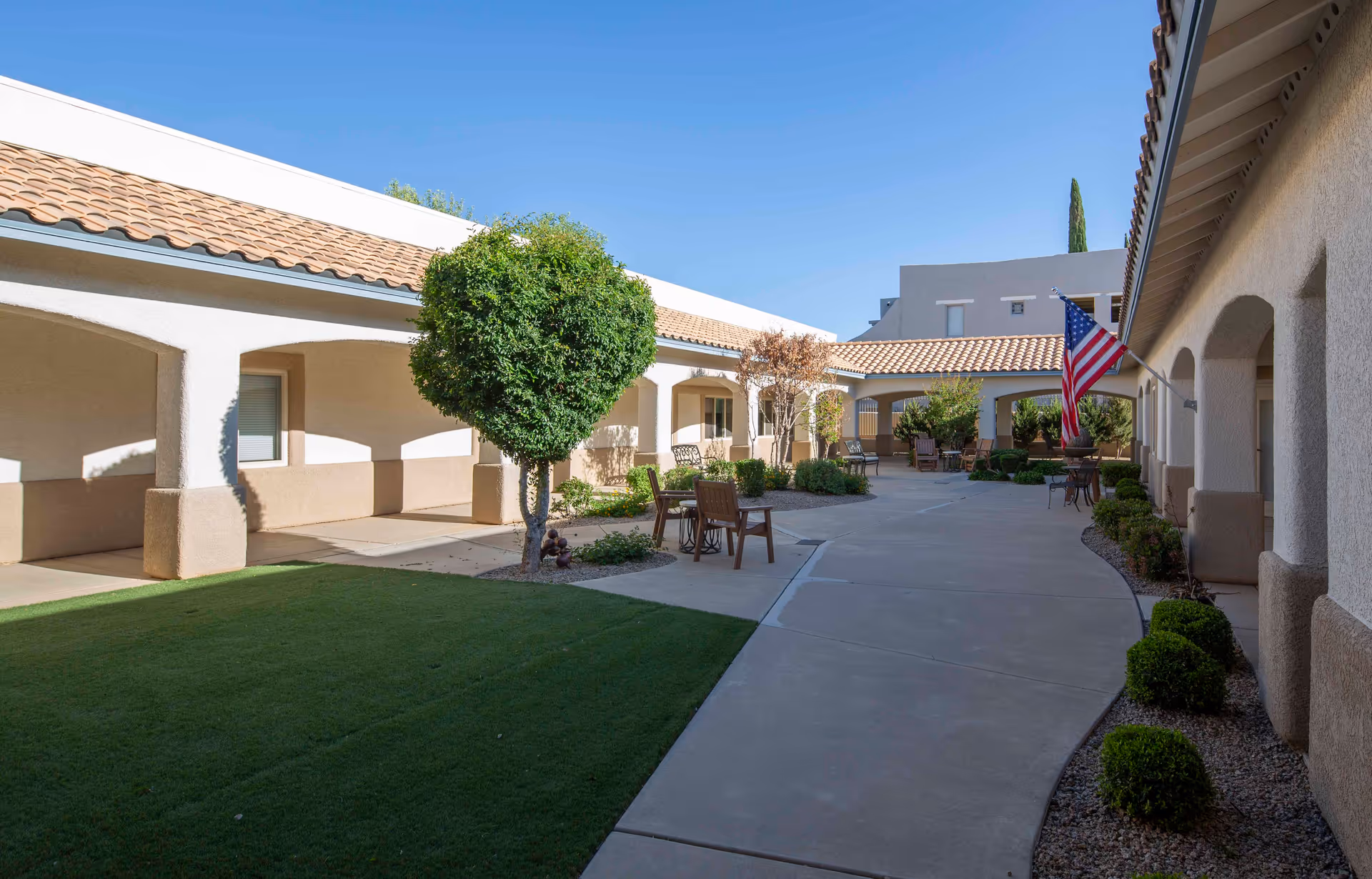 Outdoor courtyard area of Via Elegante Assisted Living Sierra Vista Highlands featuring a paved walkway, green lawn, trimmed bushes, small trees, several chairs and tables, and an American flag mounted on the building. The buildings have beige stucco walls and tiled roofs under a clear blue sky.