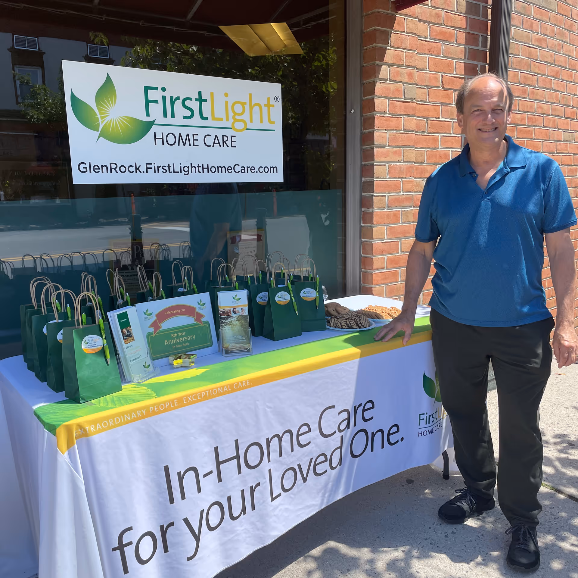 Man stands beside a promotional table with green gift bags outside FirstLight Home Care.