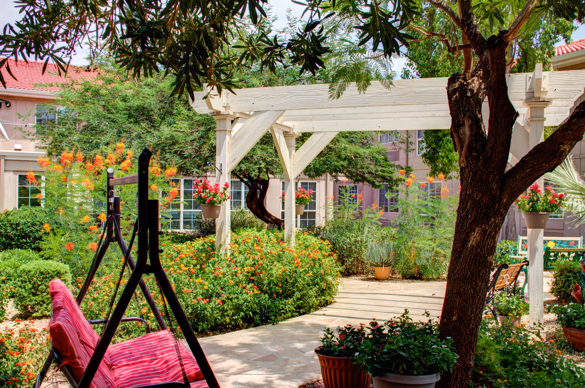 Sunlit garden courtyard with a white pergola, blooming plants, and a pink outdoor chair.