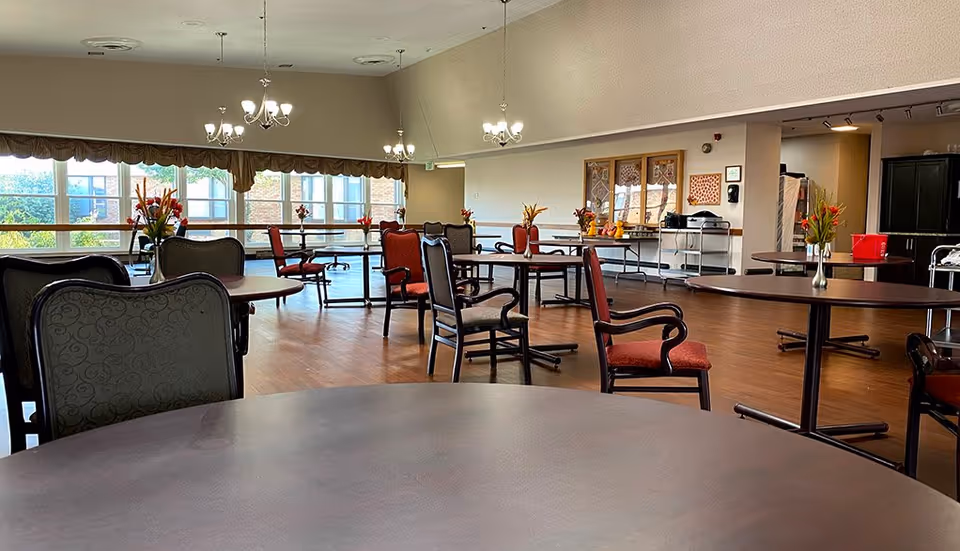 A spacious dining room with multiple round tables and chairs arranged neatly. Each table has a small vase with flowers. Large windows along one wall let in natural light, and chandeliers hang from the ceiling. The room has wooden flooring and a neutral color scheme.