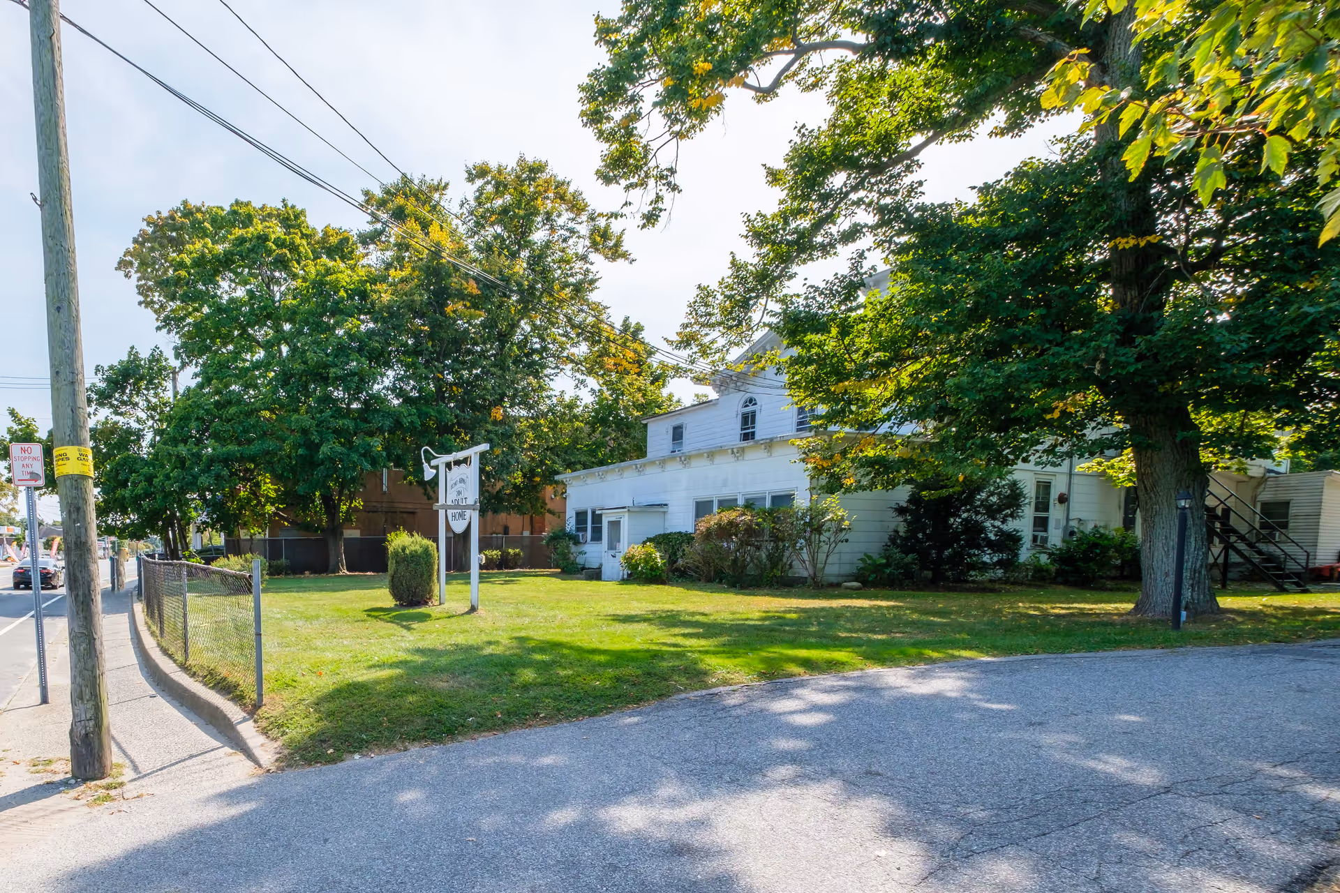 Exterior view of a white two-story building surrounded by green trees and a grassy lawn. A sign in front of the building reads 'Adult Home'. There is a sidewalk and a street with a 'No Stopping Any Time' sign on a utility pole.