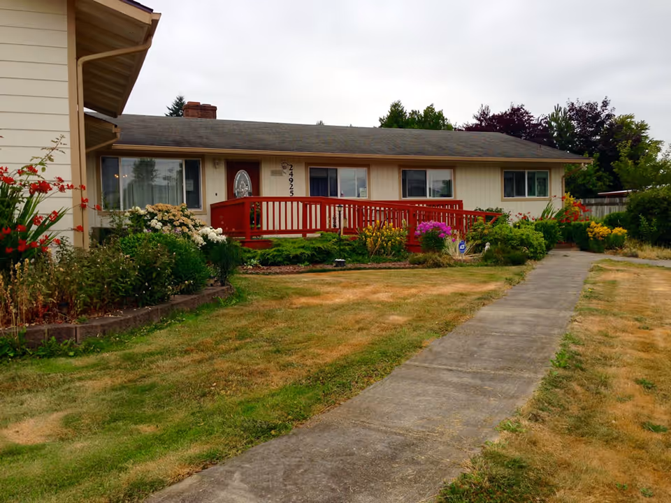 Exterior view of a single-story building with beige siding and a gray roof, featuring a red wooden ramp leading to the front door. The building is surrounded by a garden with various flowers and shrubs, and a concrete pathway runs through a lawn with patches of dry grass.