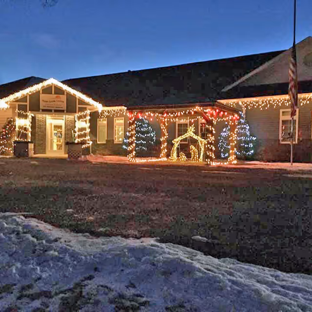 Front of Ashby Living Center decorated with Christmas lights and a lit nativity scene at dusk with snow in the foreground.