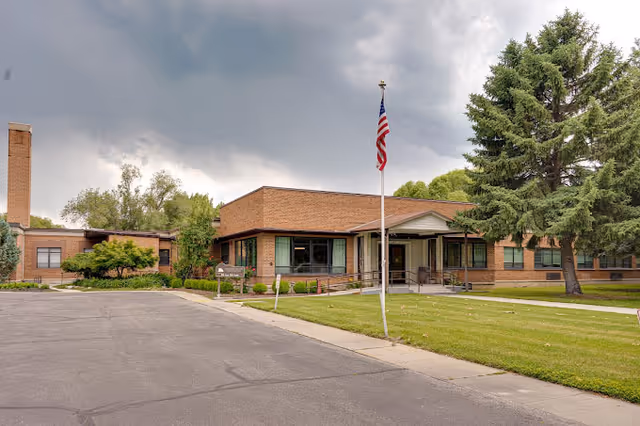 Exterior view of a single-story brick building with large windows, a covered entrance, and an American flag on a flagpole in front. The building is surrounded by green grass, trees, and a paved driveway under a cloudy sky.