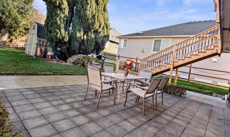 Outdoor dining patio with table and chairs near a residence, stairs, trees, and shed.