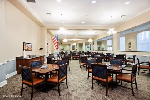 A spacious dining room with multiple wooden tables and chairs arranged neatly. The room has beige walls, carpeted flooring, and several chandeliers hanging from the ceiling. An American flag is displayed on the left side near the wall, and there are framed pictures on the walls. The room is well-lit with natural light coming through windows on the right side.