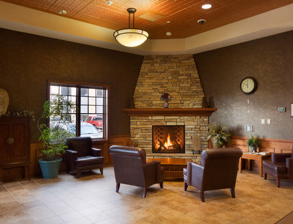Cozy lounge with a stone fireplace flanked by leather armchairs, a window, and potted plants.
