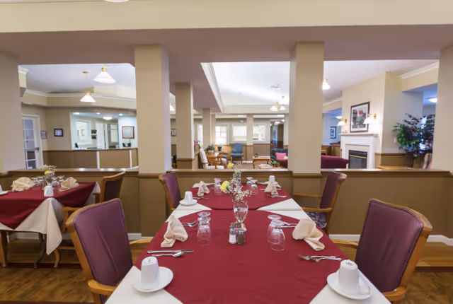Dining area in a senior living facility with tables covered in burgundy and beige tablecloths, set with cups, glasses, napkins, and silverware. The room has wooden floors, beige walls, and is divided by half walls and columns. In the background, there is a living room area with chairs, a fireplace, and plants.