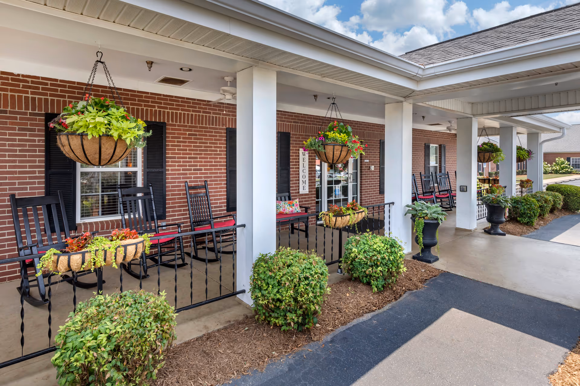 Covered outdoor porch area with hanging flower baskets, black rocking chairs with red cushions, and green shrubs along the edge. The building has red brick walls and white columns supporting the roof. A welcome sign is visible near the entrance door.