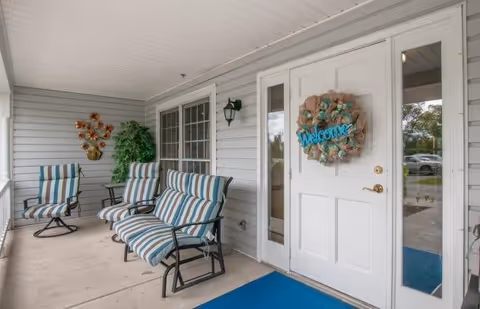 Covered front porch with a white door decorated with a "Welcome" wreath, three striped cushioned chairs, potted plants, and a blue doormat.
