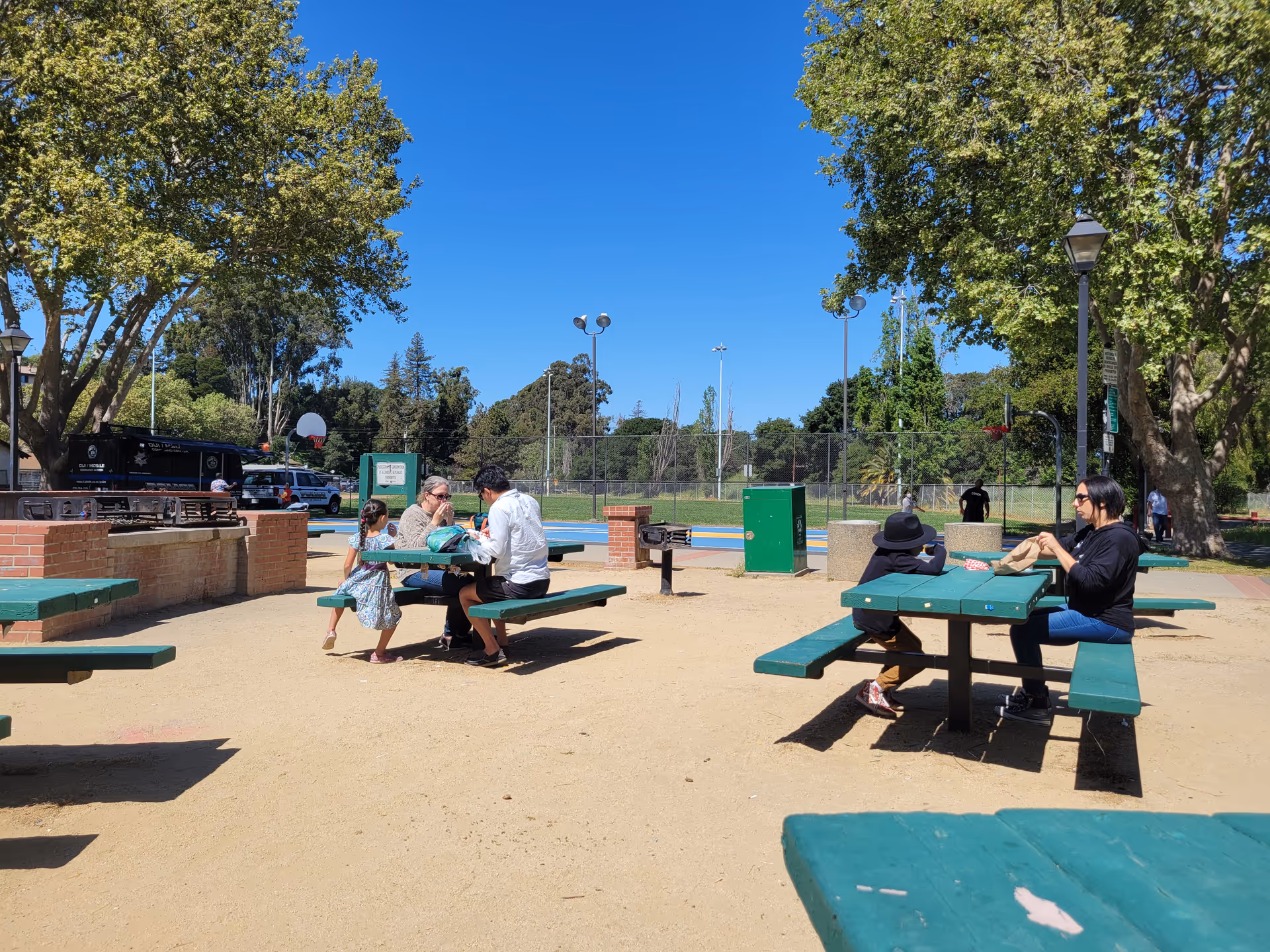 Sunny outdoor park area with green picnic tables and people sitting and chatting.
