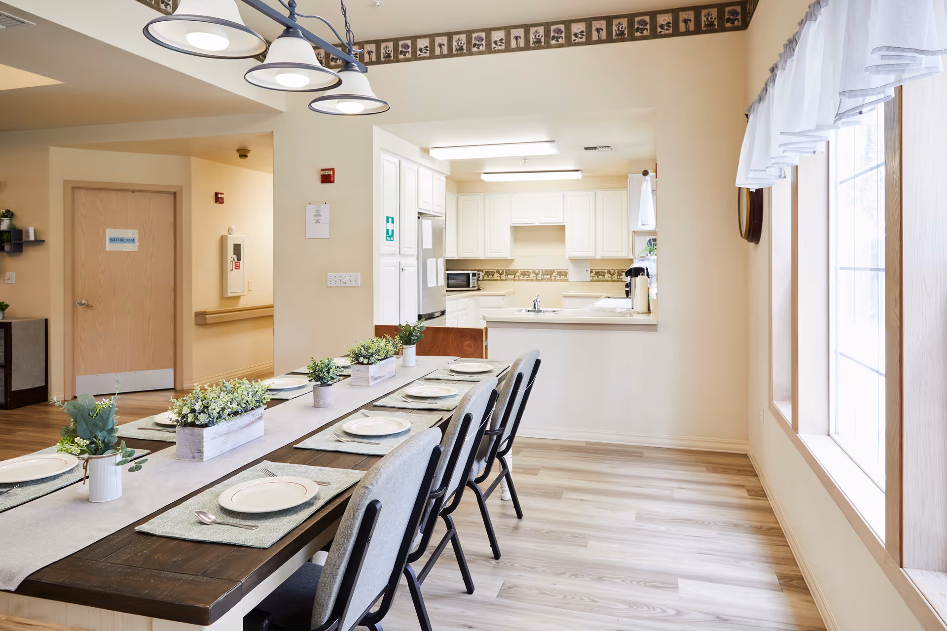 A bright dining area with a long wooden table set with plates, placemats, and small potted plants as centerpieces. The room has light-colored walls and flooring, a large window with sheer white curtains on the right, and a view into a white kitchen with cabinets and appliances. A door labeled 'Bathroom' is visible in the background.