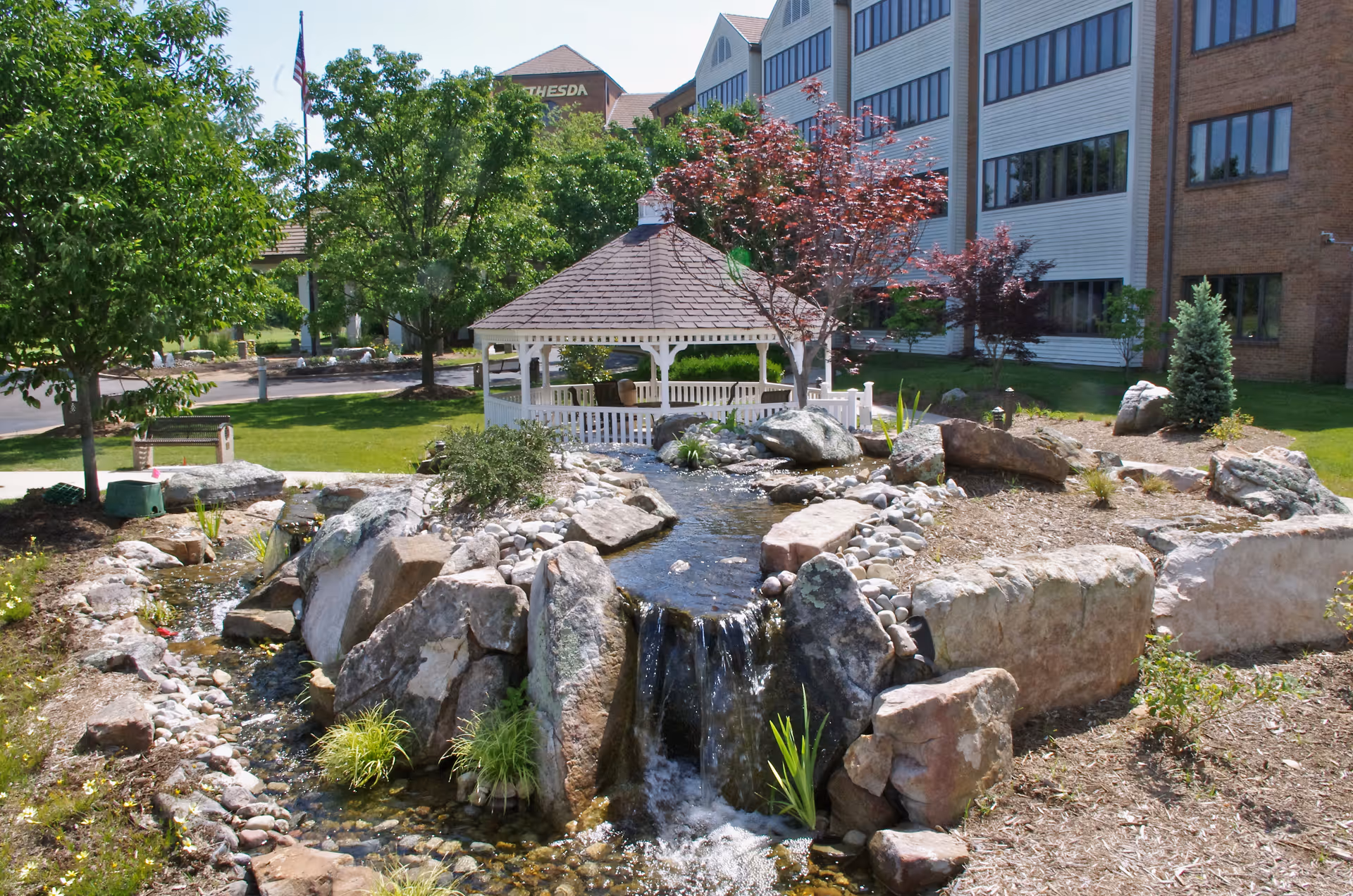 Outdoor garden area at Ellisville Rehab and Nursing featuring a small waterfall flowing over rocks into a pond, a white gazebo surrounded by trees and shrubs, and a multi-story building in the background.
